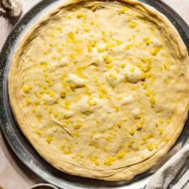 Unbaked pizza dough on a round metal tray, brushed with garlic butter. Fresh thyme, garlic, a pastry brush, and a striped towel are nearby.