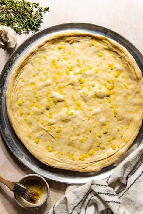 Unbaked pizza dough on a round metal tray, brushed with garlic butter. Fresh thyme, garlic, a pastry brush, and a striped towel are nearby.