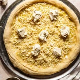 Unbaked pizza dough topped with shredded cheese and dollops of ricotta on a round metal tray, with a pastry brush, striped towel, herbs, and garlic nearby.