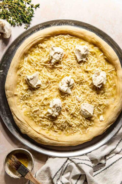 Unbaked pizza dough topped with shredded cheese and dollops of ricotta on a round metal tray, with a pastry brush, striped towel, herbs, and garlic nearby.
