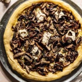 A pizza topped with sautéed mushrooms and dollops of ricotta cheese on a metal tray, with fresh thyme and garlic nearby.