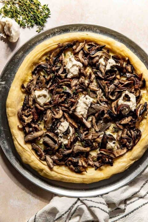 A pizza topped with sautéed mushrooms and dollops of ricotta cheese on a metal tray, with fresh thyme and garlic nearby.