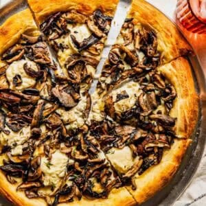 A sliced mushroom and cheese pizza on a round metal tray, with a pizza cutter, a glass of iced drink, and a striped napkin nearby.