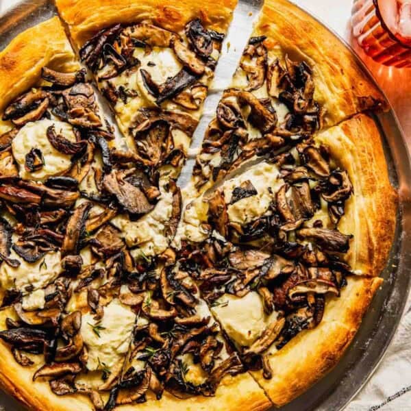 A sliced mushroom and cheese pizza on a round metal tray, with a pizza cutter, a glass of iced drink, and a striped napkin nearby.