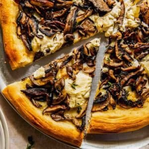 A close-up of a sliced pizza topped with ricotta cheese and sautéed mushrooms on a round metal tray.