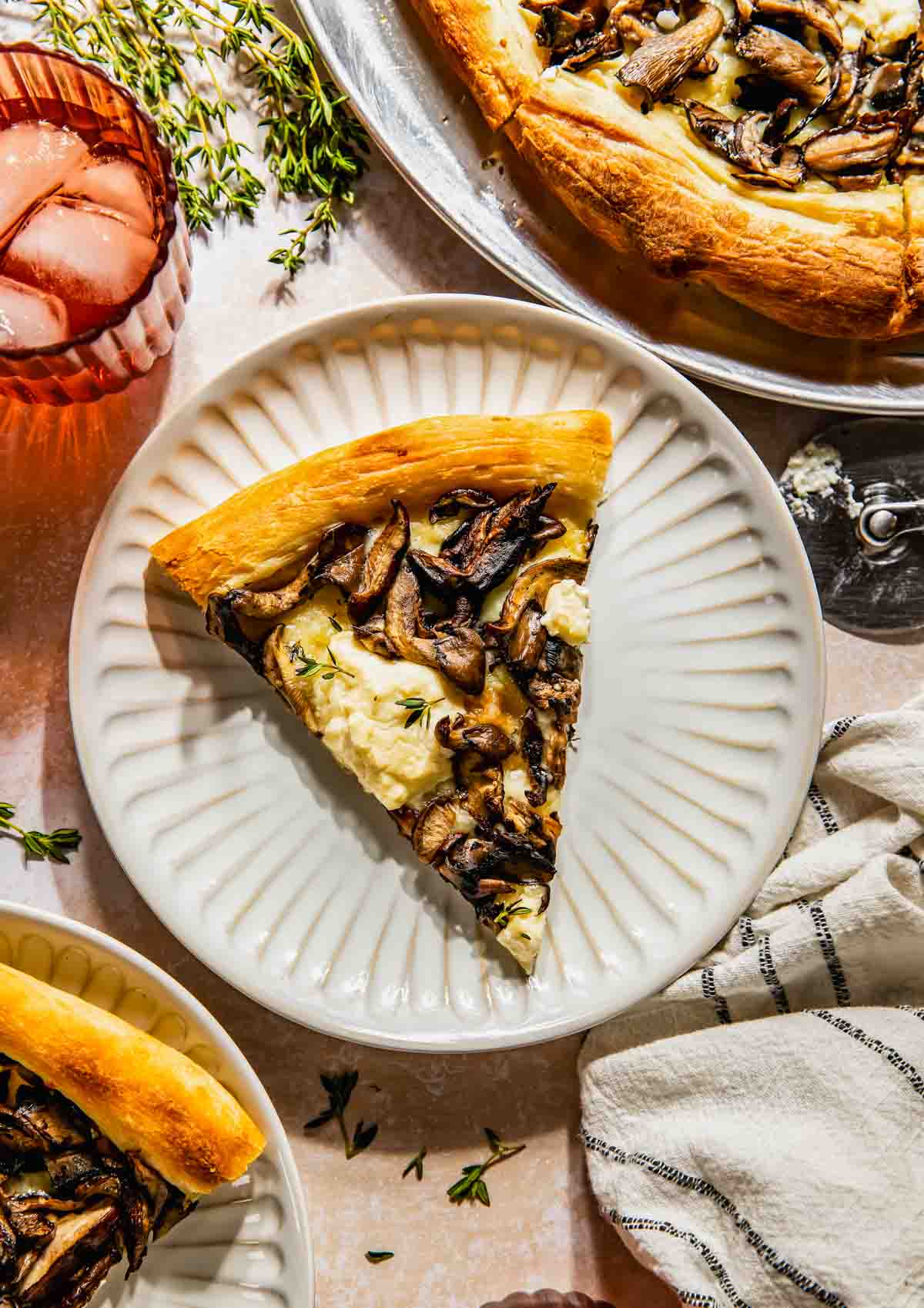 Ein Stück Pilz-Ricotta-Pizza liegt auf einem weißen Teller, umgeben von einer Serviette, Kräutern, einem Pizzaschneider, einem Glas Eis und einem rosa Getränk.