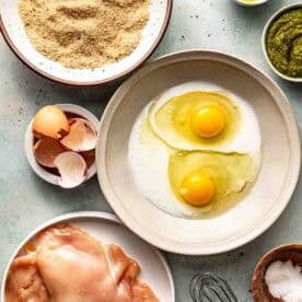 A flat lay of raw chicken breasts, eggs with milk in a bowl, bread crumbs, pesto, olive oil, egg shells, salt, and a whisk on a light surface.