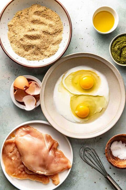 A flat lay of raw chicken breasts, eggs with milk in a bowl, bread crumbs, pesto, olive oil, egg shells, salt, and a whisk on a light surface.