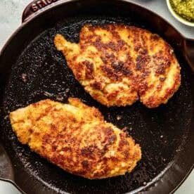 Two breaded and pan-fried chicken breasts cooking in a black skillet, with small bowls of salt, pesto, and cheese nearby.