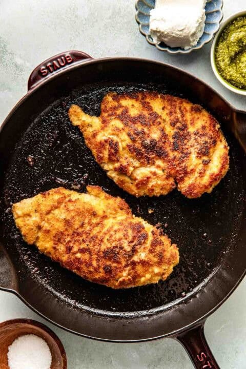 Two breaded and pan-fried chicken breasts cooking in a black skillet, with small bowls of salt, pesto, and cheese nearby.