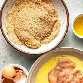 Two bowls: one with raw chicken in beaten egg, the other with chicken coated in breadcrumbs. Eggshells and a small bowl of melted butter are nearby.