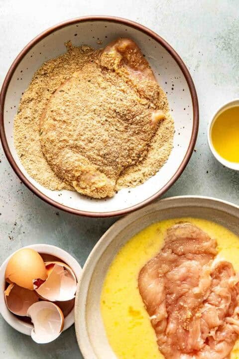 Two bowls: one with raw chicken in beaten egg, the other with chicken coated in breadcrumbs. Eggshells and a small bowl of melted butter are nearby.