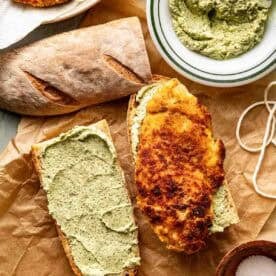 A sandwich being assembled with crusty bread, a layer of pesto, and a piece of breaded, fried cutlet, alongside extra bread, pesto spread, and a small bowl of salt.