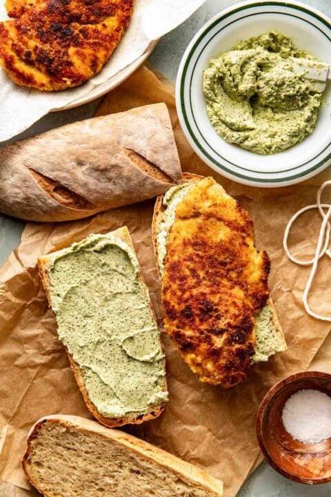A sandwich being assembled with crusty bread, a layer of pesto, and a piece of breaded, fried cutlet, alongside extra bread, pesto spread, and a small bowl of salt.