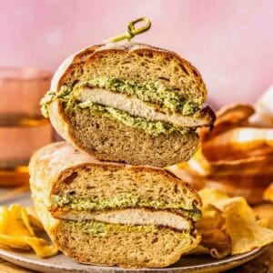 A chicken sandwich with pesto sauce is cut in half and stacked on a plate, surrounded by potato chips.
