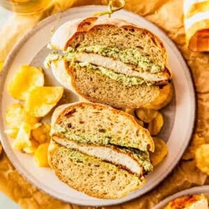 A sandwich with breaded chicken and green pesto filling is cut in half on a plate, surrounded by potato chips and a drink.