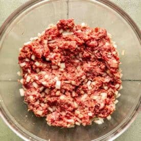 A glass bowl containing a mixture of raw ground meat and diced onions on a gray tiled surface.