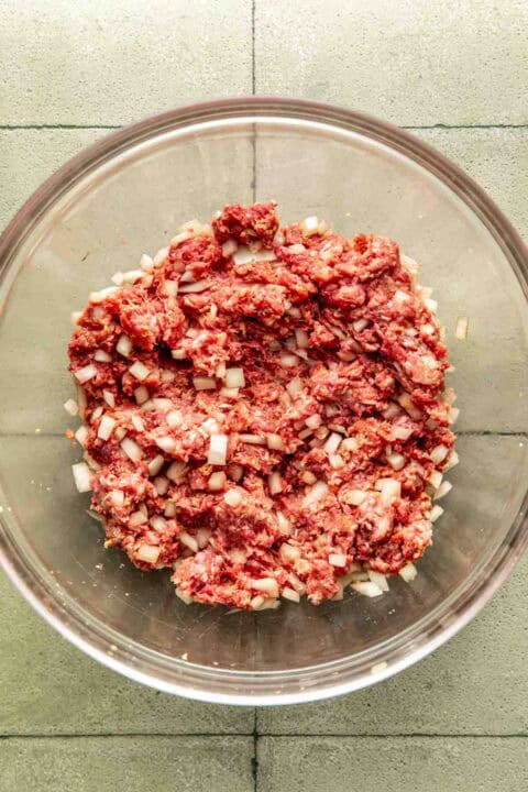 A glass bowl containing a mixture of raw ground meat and diced onions on a gray tiled surface.