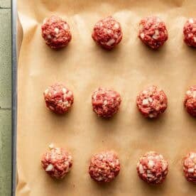 Uncooked meatballs with chopped onions are arranged in rows on a parchment-lined baking sheet.