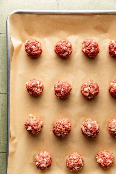 Uncooked meatballs with chopped onions are arranged in rows on a parchment-lined baking sheet.