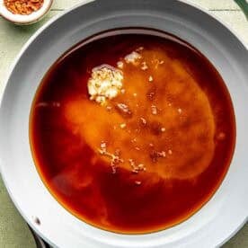 A white skillet with soy sauce, minced garlic, and brown sugar mixture; a small bowl of red pepper flakes sits beside the pan on a tiled surface.