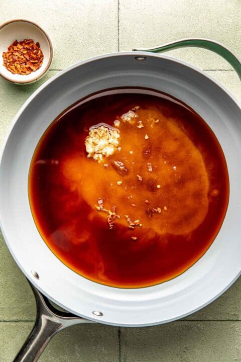 A white skillet with soy sauce, minced garlic, and brown sugar mixture; a small bowl of red pepper flakes sits beside the pan on a tiled surface.