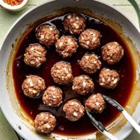 A pan of uncooked meatballs with chopped onions sits in a dark sauce. Metal tongs rest on the pans edge. A small bowl of red pepper flakes is nearby.