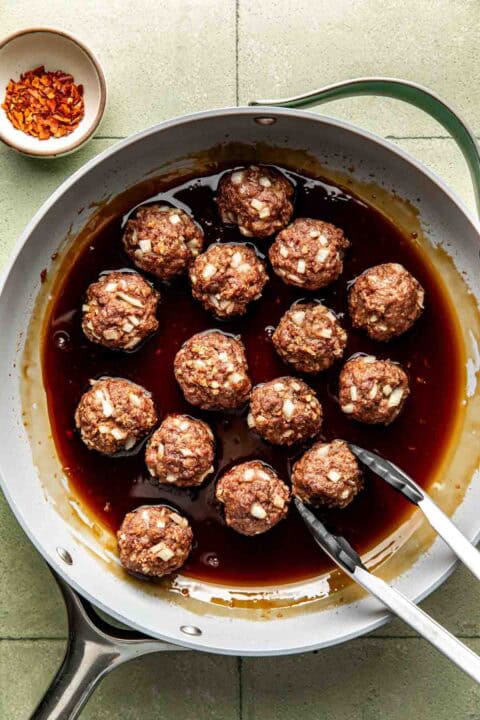 A pan of uncooked meatballs with chopped onions sits in a dark sauce. Metal tongs rest on the pans edge. A small bowl of red pepper flakes is nearby.