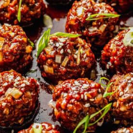 Close-up of glazed meatballs topped with sesame seeds and sliced green onions, arranged on a dark sauce.