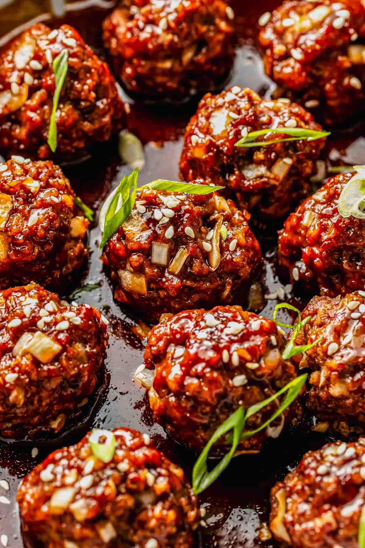 Close-up of glazed meatballs topped with sesame seeds and sliced green onions, arranged on a dark sauce.