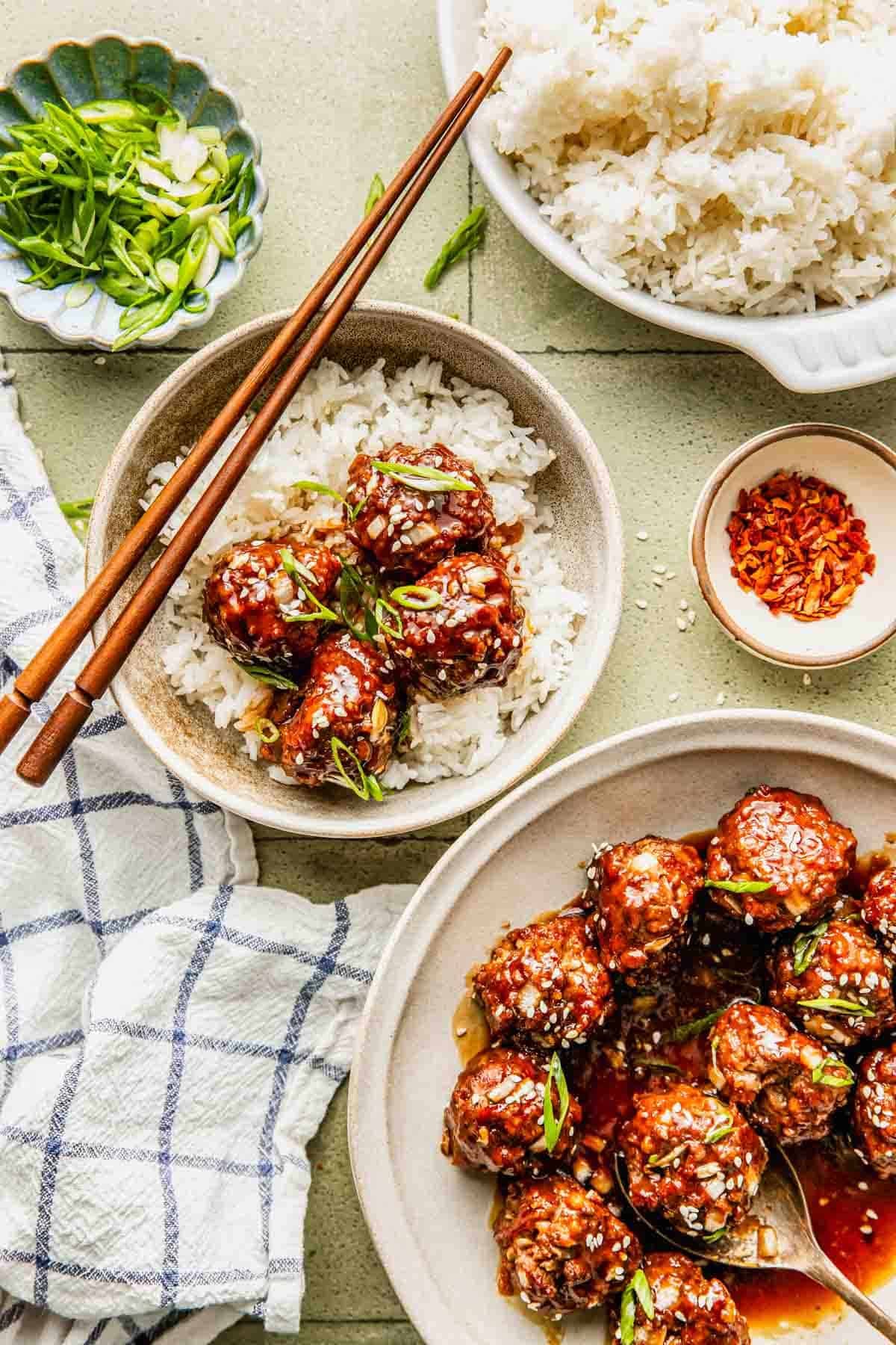 A bowl of white rice topped with glazed meatballs, garnished with sesame seeds and green onions; chopsticks rest on the bowl. Other dishes of rice, meatballs, and seasonings are nearby.