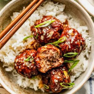A bowl of white rice topped with glazed meatballs, garnished with sesame seeds and sliced green onions, with chopsticks resting on the bowl.