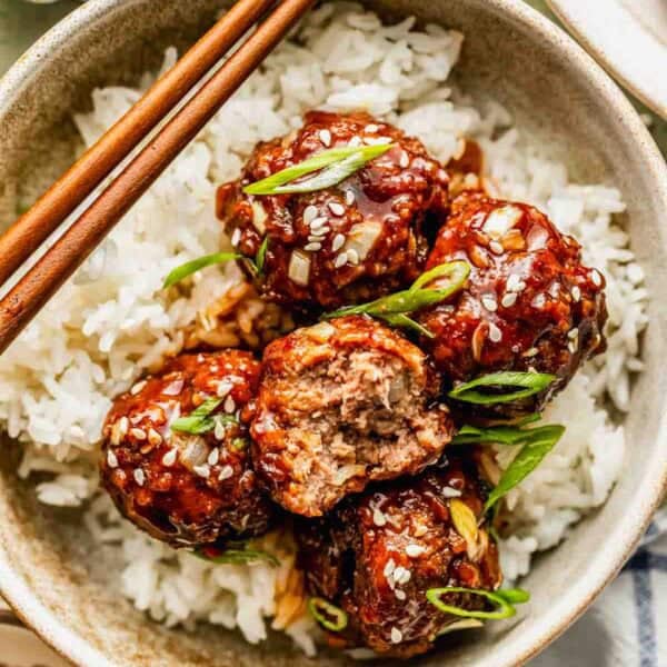 A bowl of white rice topped with glazed meatballs, garnished with sesame seeds and sliced green onions, with chopsticks resting on the bowl.