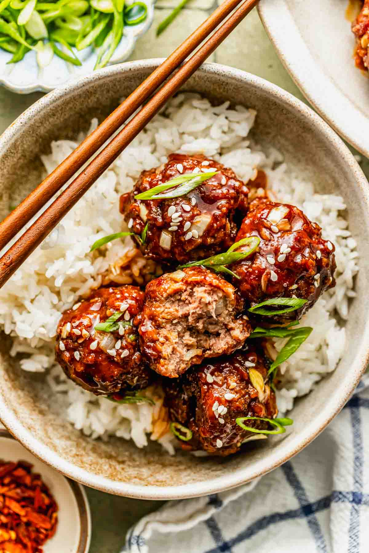 A bowl of white rice topped with glazed meatballs, garnished with sesame seeds and sliced green onions, with chopsticks resting on the bowl.