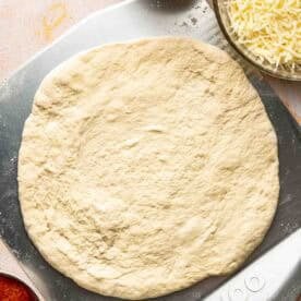 Unbaked pizza dough on a metal pizza peel, surrounded by bowls of tomato sauce, shredded cheese, sliced pepperoni, and cooked sausage.