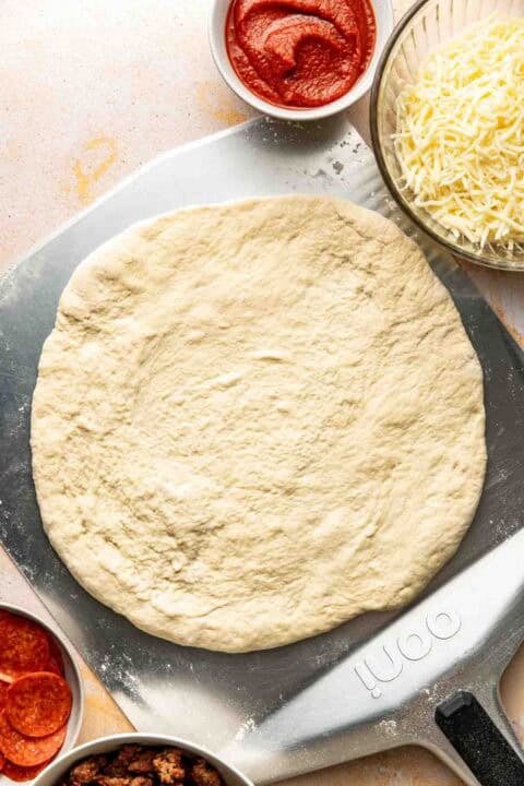 Unbaked pizza dough on a metal pizza peel, surrounded by bowls of tomato sauce, shredded cheese, sliced pepperoni, and cooked sausage.