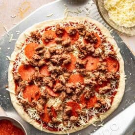 Unbaked pizza on a metal peel topped with tomato sauce, shredded cheese, pepperoni, and cooked sausage, surrounded by bowls of sauce, cheese, pepperoni, and sausage.