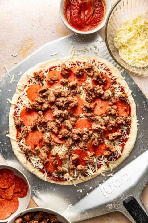 Unbaked pizza on a metal peel topped with tomato sauce, shredded cheese, pepperoni, and cooked sausage, surrounded by bowls of sauce, cheese, pepperoni, and sausage.
