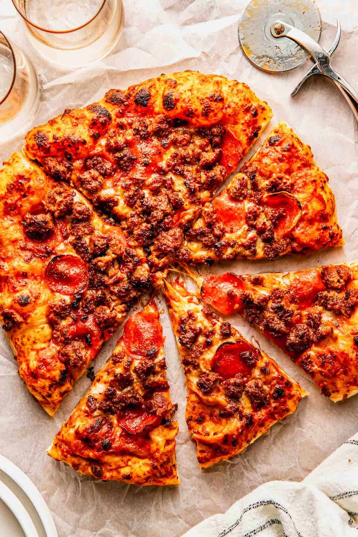 A sliced sausage and pepperoni pizza on parchment paper, with a pizza cutter and two drinking glasses nearby.