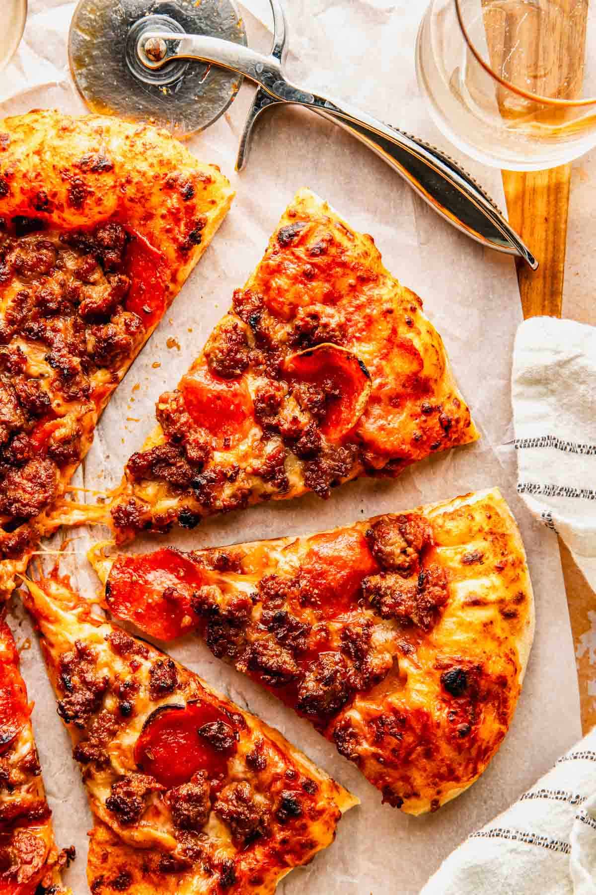 Overhead view of sliced pepperoni and sausage pizza on parchment paper, with a pizza cutter and a glass nearby.