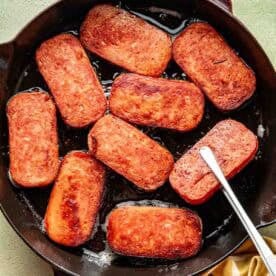 Slices of Spam are being pan-fried in a black cast iron skillet, with a metal spatula and a yellow checkered towel beside the pan.