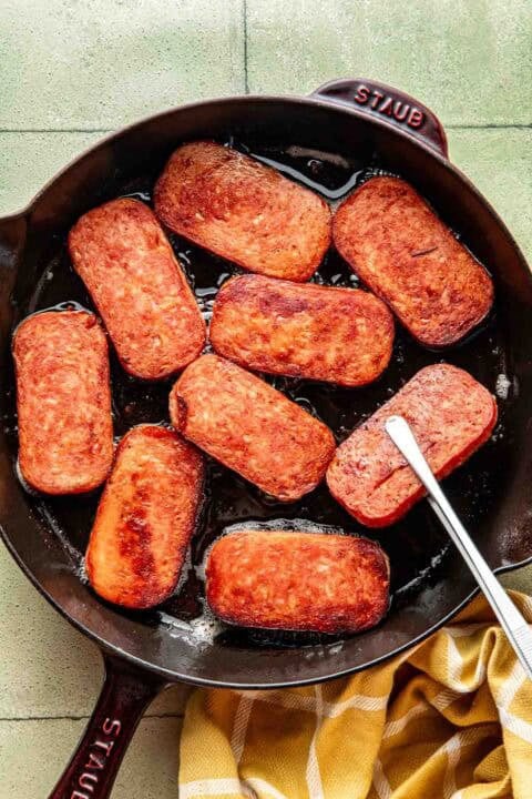 Slices of Spam are being pan-fried in a black cast iron skillet, with a metal spatula and a yellow checkered towel beside the pan.