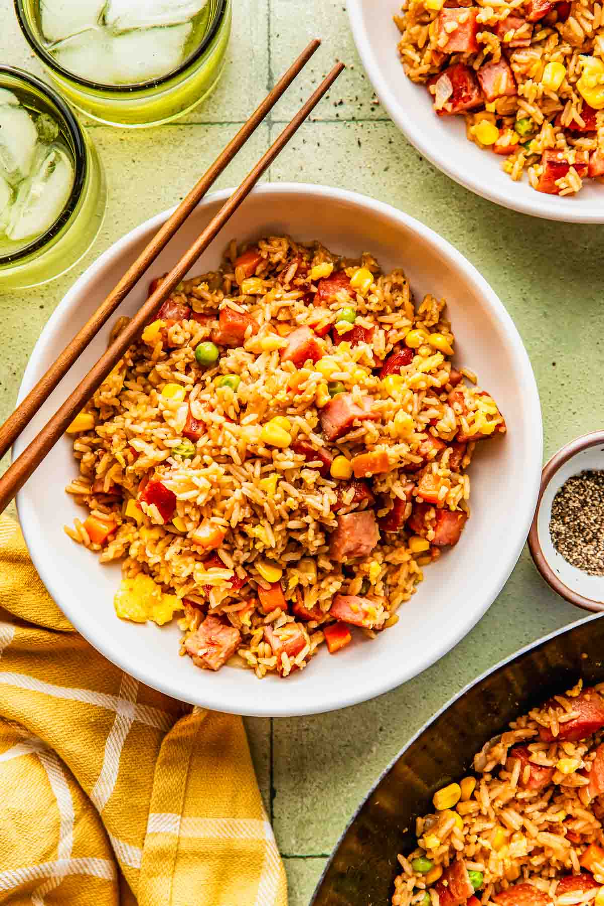 A bowl of fried rice with vegetables and diced meat, served with chopsticks, ice water, and a small bowl of pepper on a light green surface.