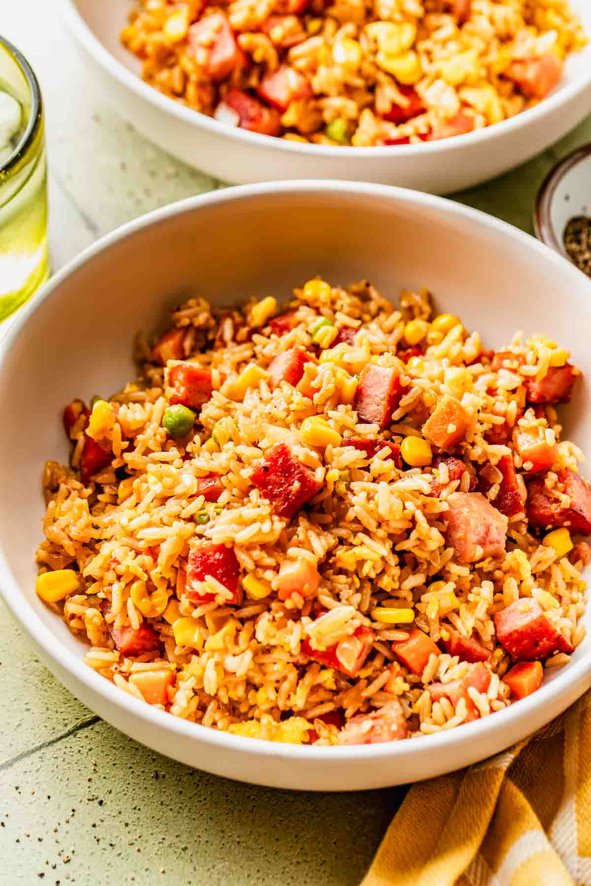 A white bowl filled with fried rice mixed with diced ham, corn, carrots, and peas. A second bowl of the same dish is partially visible in the background.