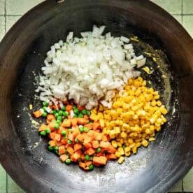 Chopped onions, diced carrots with peas, and corn kernels are arranged in a wok on a tiled surface, ready to be cooked.