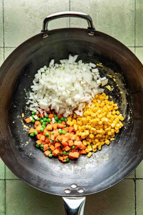 Chopped onions, diced carrots with peas, and corn kernels are arranged in a wok on a tiled surface, ready to be cooked.