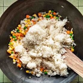 A wok with cooked white rice, mixed vegetables, and a wooden spatula on a green tiled surface, ready for stir-frying.