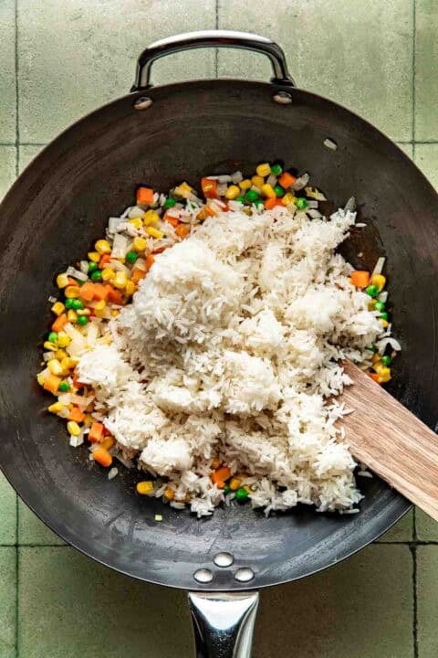 A wok with cooked white rice, mixed vegetables, and a wooden spatula on a green tiled surface, ready for stir-frying.