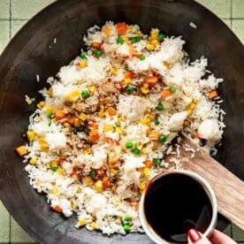A hand holds a bowl of soy sauce over a wok containing rice, mixed vegetables, and tuna on a tiled surface.