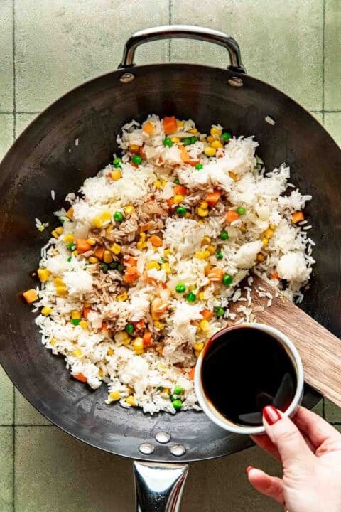 A hand holds a bowl of soy sauce over a wok containing rice, mixed vegetables, and tuna on a tiled surface.